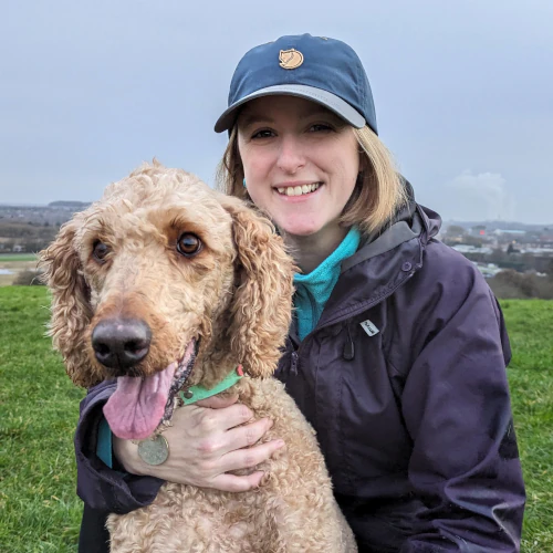 Rebecca smiling outdoors while holding Omar, a goldendoodle, on a grassy hillside.