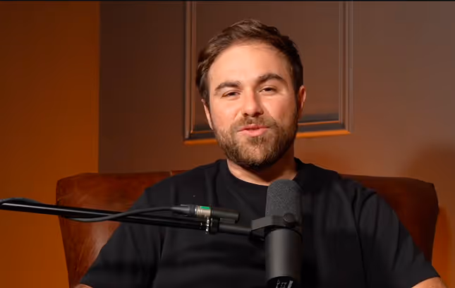 Un homme souriant avec une barbe assis devant un microphone dans un cadre intérieur aux tons chauds.