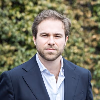 Portrait d'un homme aux cheveux bruns courts et barbe légère, portant un costume bleu foncé et une chemise blanche, avec un arrière-plan de feuillage flou.