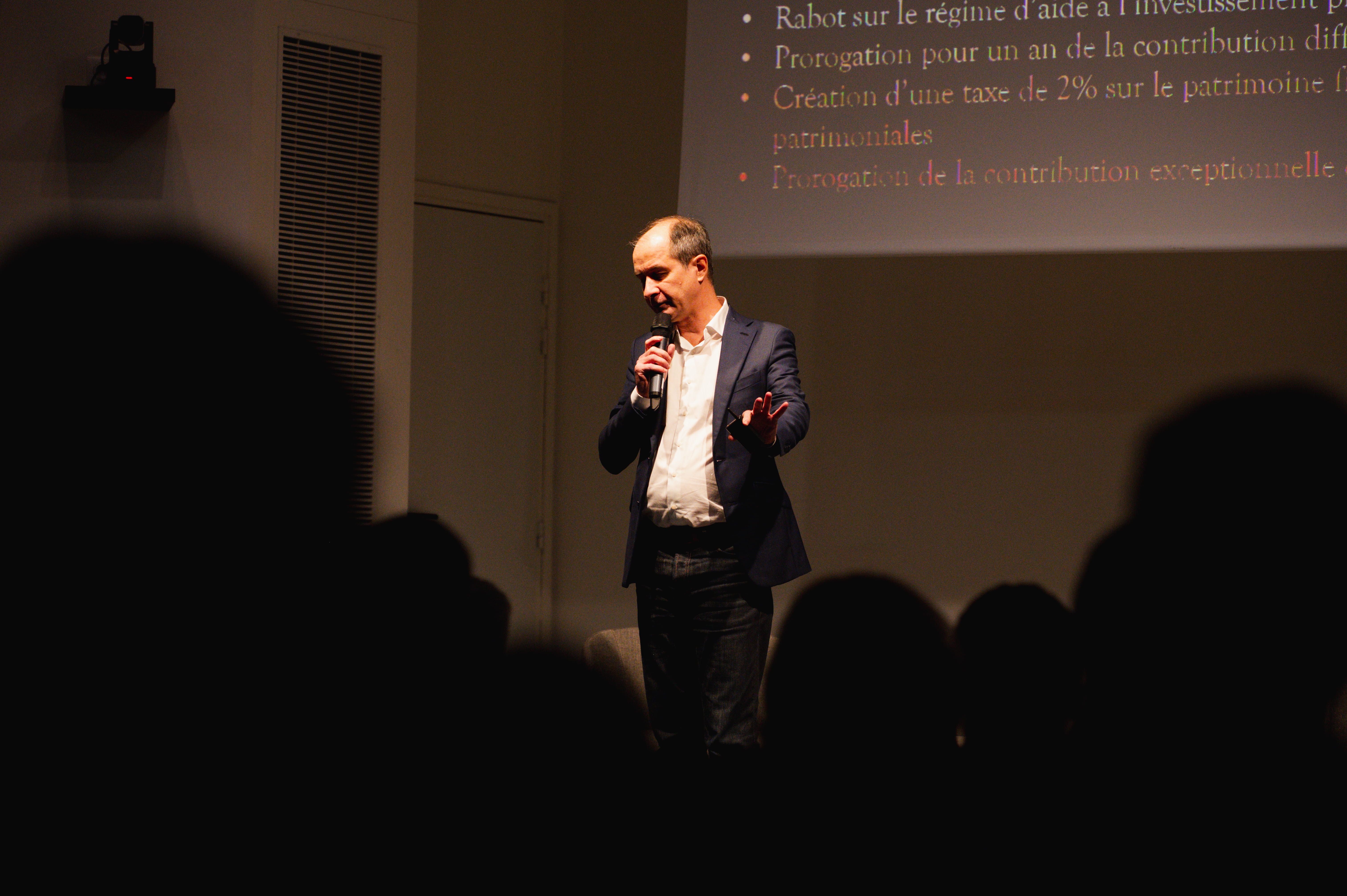 Un homme tient un micro et parle devant un public avec un écran de présentation projeté en arrière-plan.