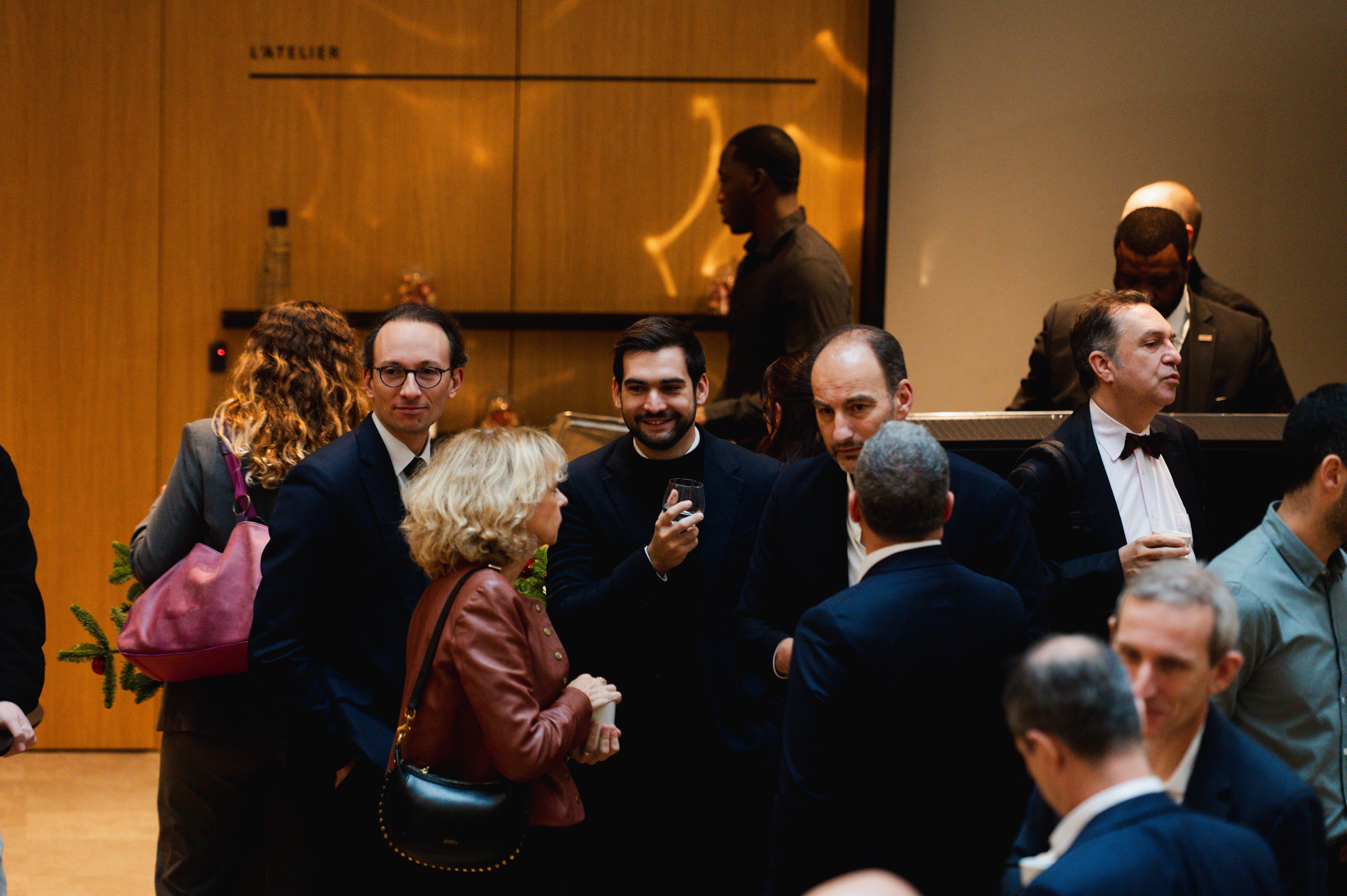 Un groupe de personnes habillées formellement discutant lors d'un événement social en intérieur avec un mur en bois en arrière-plan.