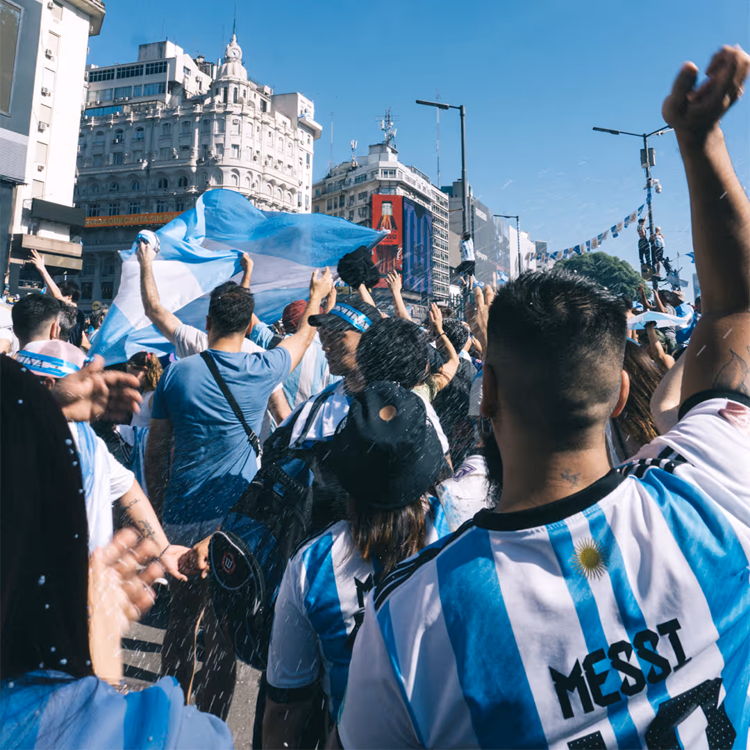 Crowd celebrating outdoors waving Argentine flags and wearing Argentina jerseys under a clear blue sky.