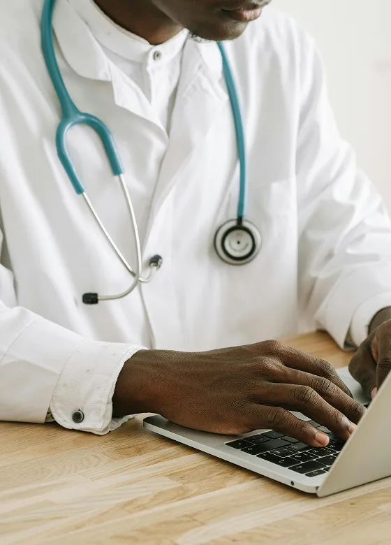 Doctor in white coat with stethoscope typing on laptop at desk.