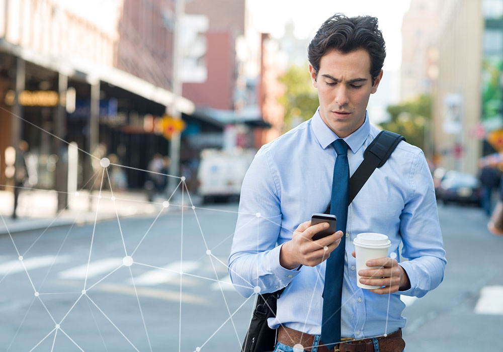 Businessman using smartphone while holding coffee cup on city street with digital network overlay graphics