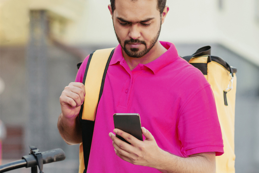 Food delivery worker in pink shirt checking phone while carrying yellow delivery bag, illustrating food delivery problems