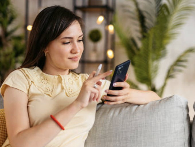 Woman sitting on a couch, holding and using a smartphone with a stylus pen indoors.