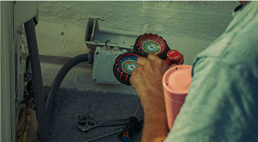 Person holding a manifold gauge set near HVAC unit pipes against a wall.