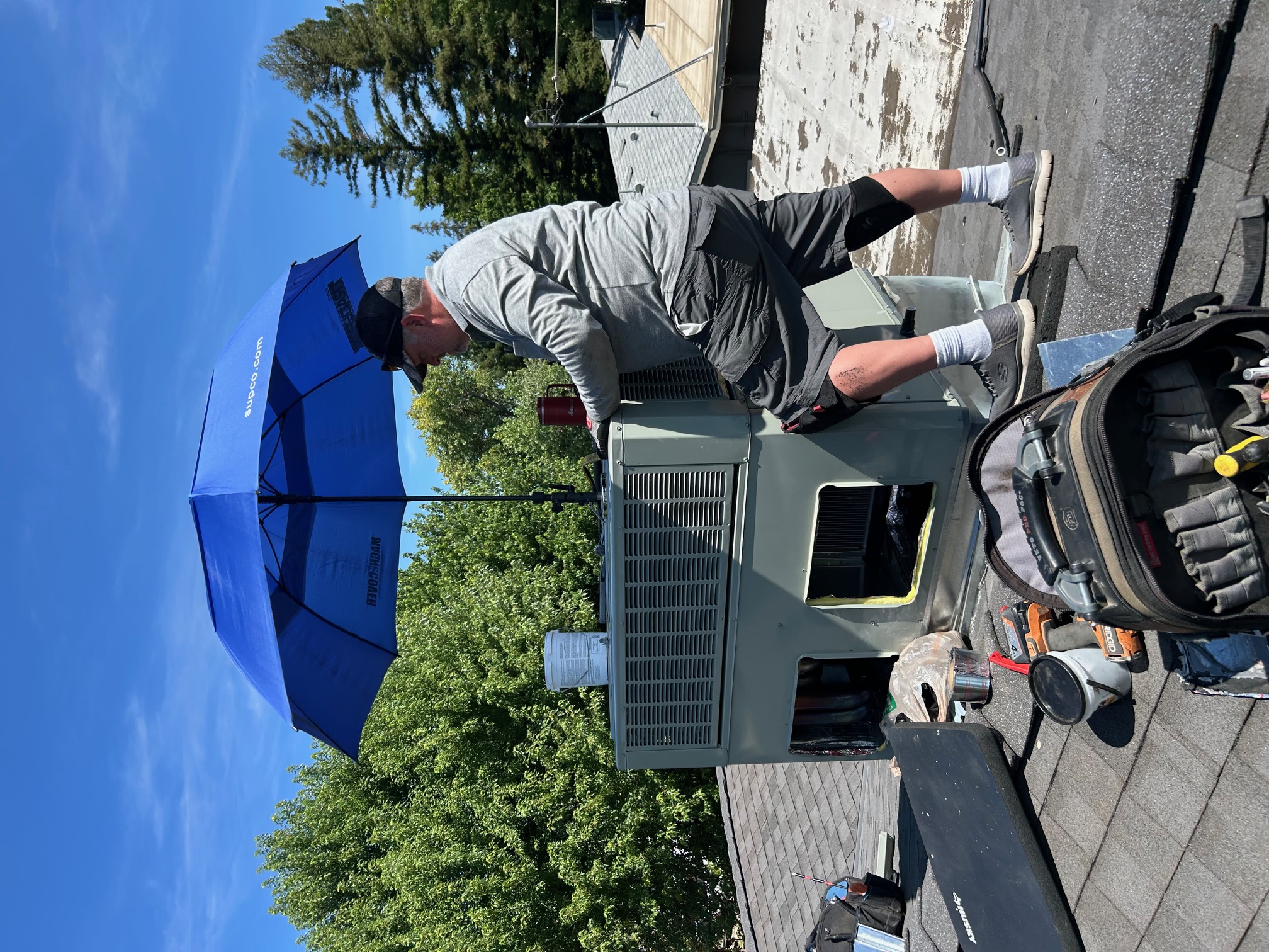 Technician working on a rooftop HVAC unit under a blue umbrella with tools scattered around.