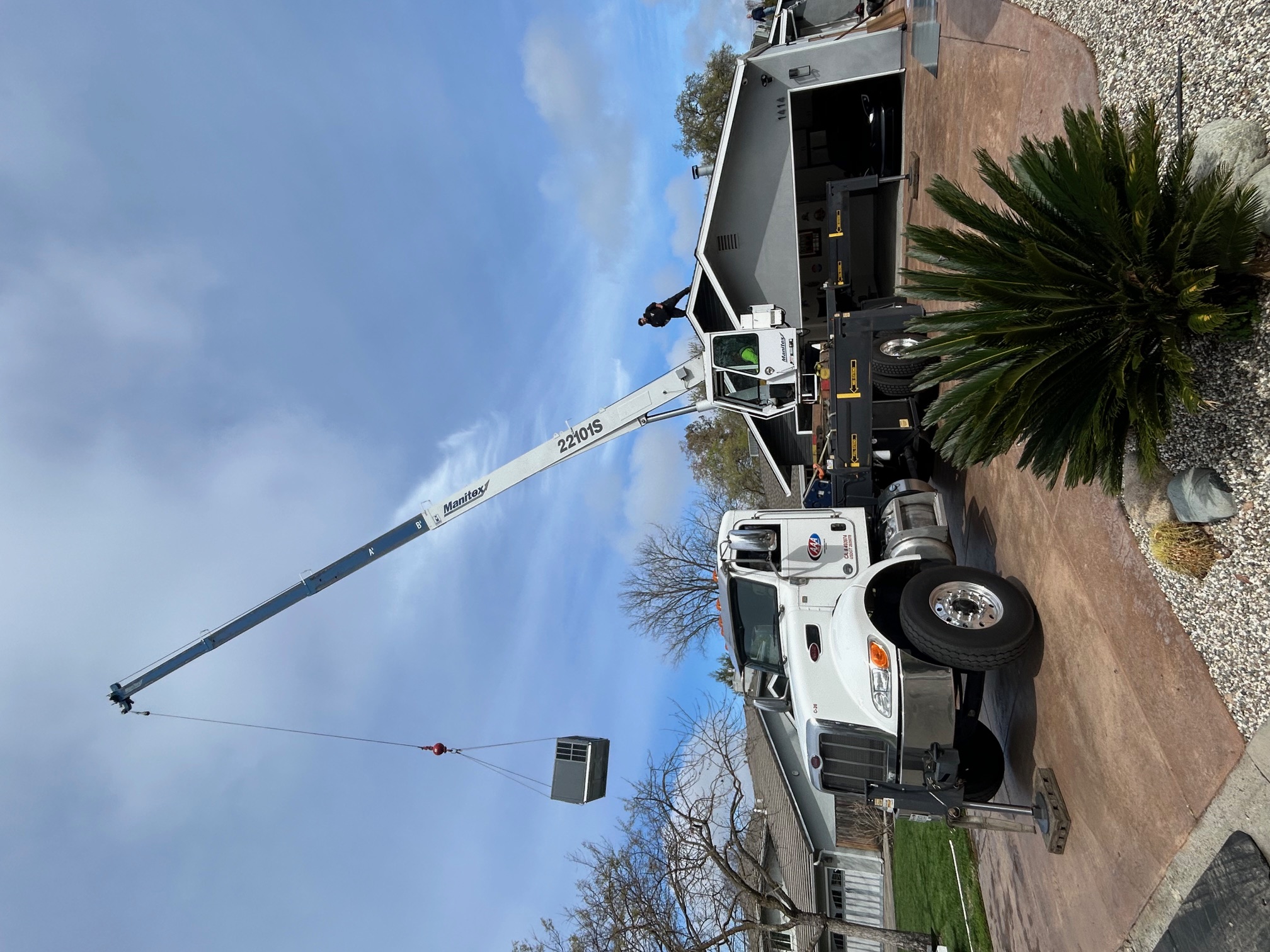 White truck-mounted crane lifting a large HVAC unit with two people working on a residential rooftop.