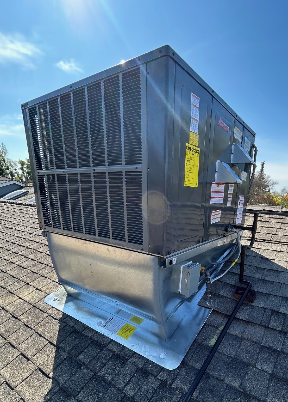 Large HVAC packaged unit installed on a shingled rooftop under a clear blue sky with sunlight flare.