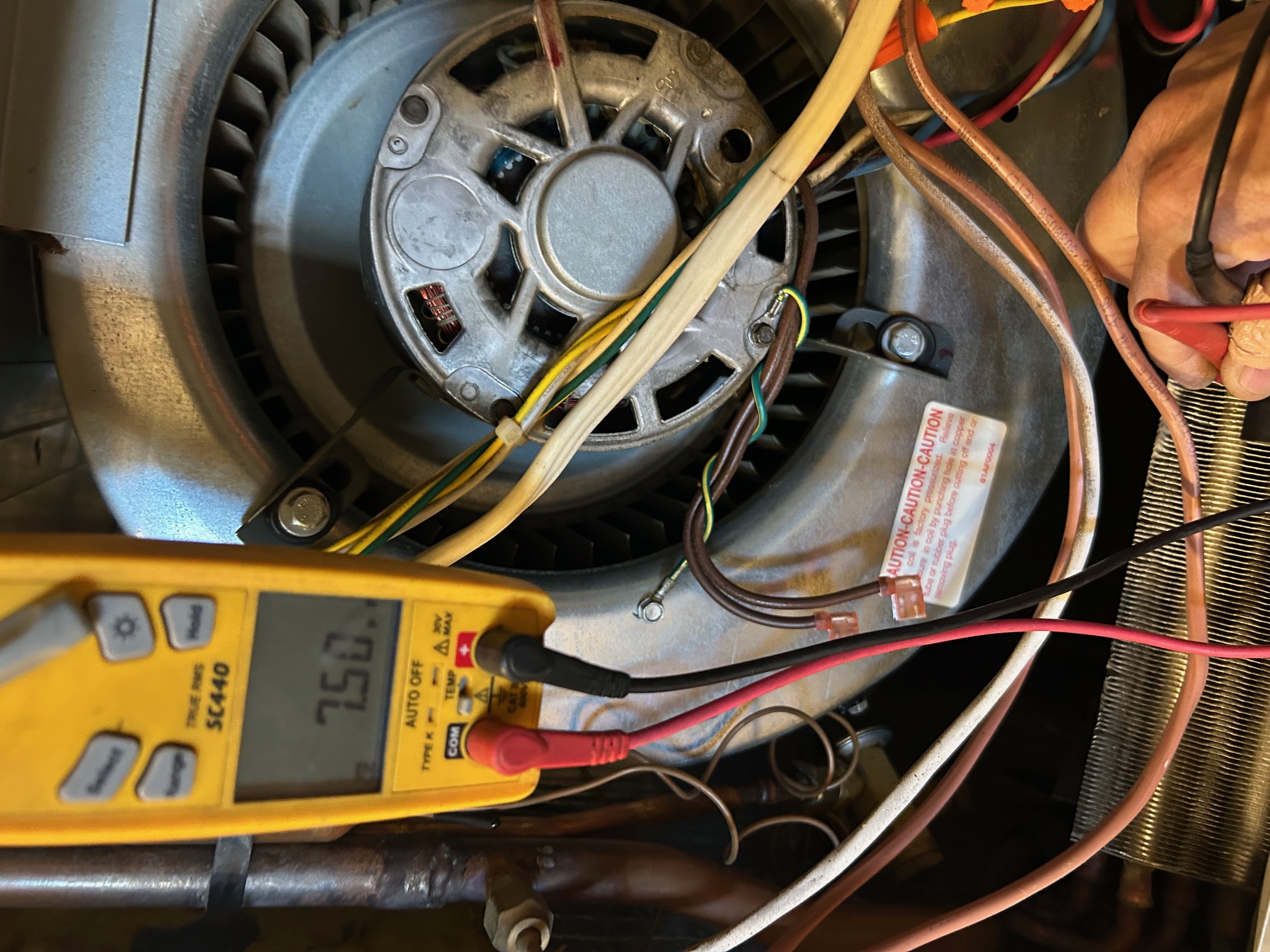 Hand holding red probe connected to a yellow digital multimeter displaying 7.50, testing wires near a metal fan inside an electrical device.