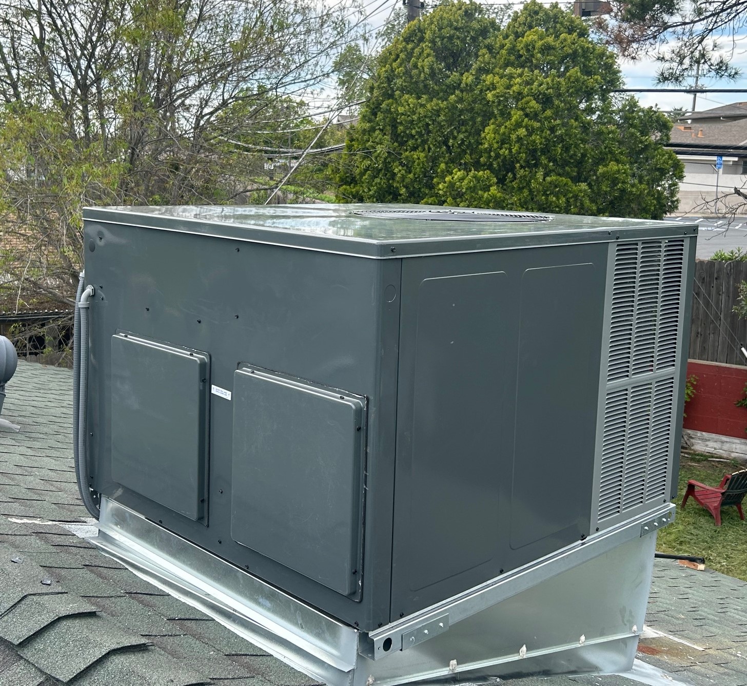 Gray rooftop packaged HVAC unit installed on shingled roof with trees and a fence in the background.