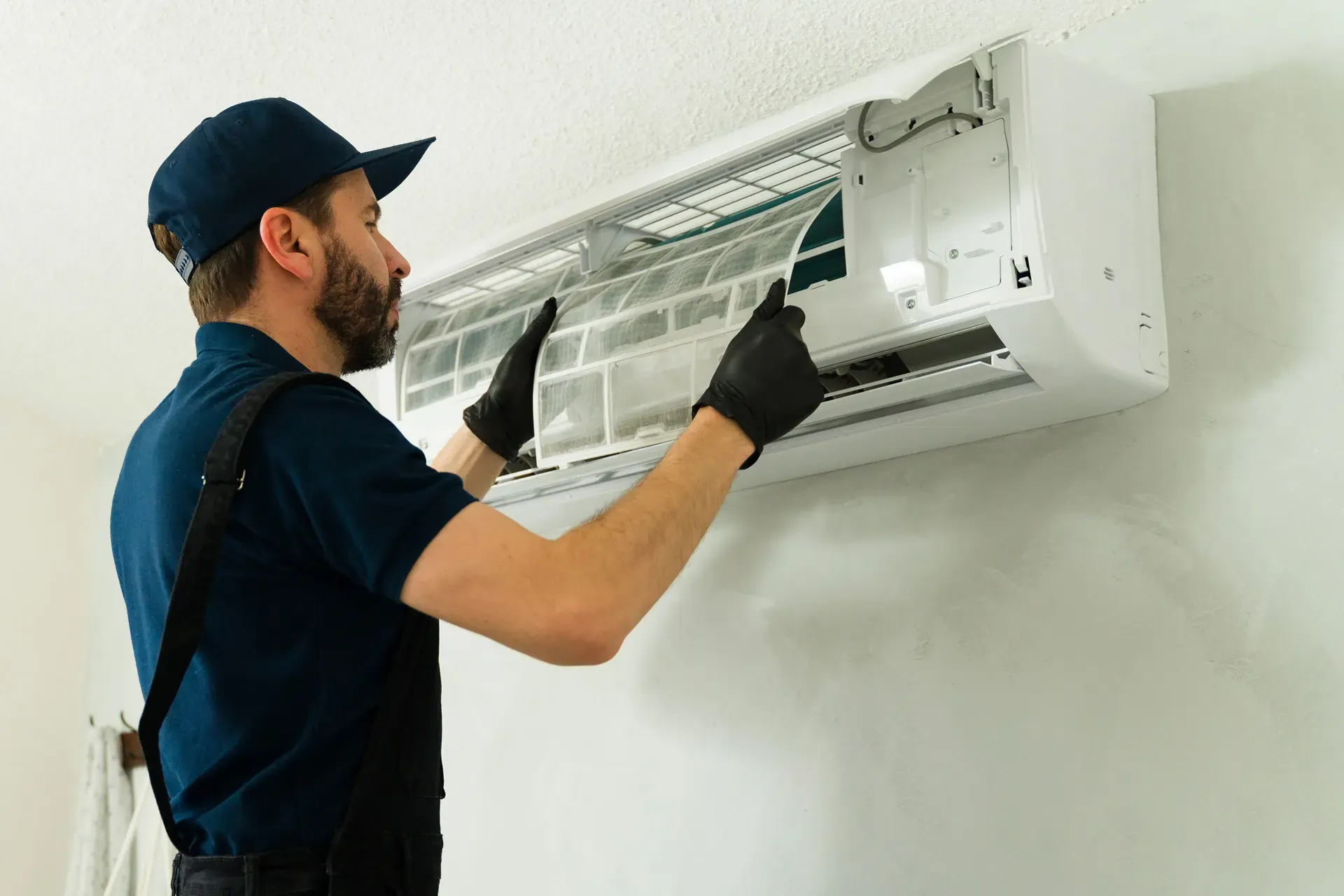 Technician wearing black gloves inspecting and cleaning a wall-mounted air conditioner unit.