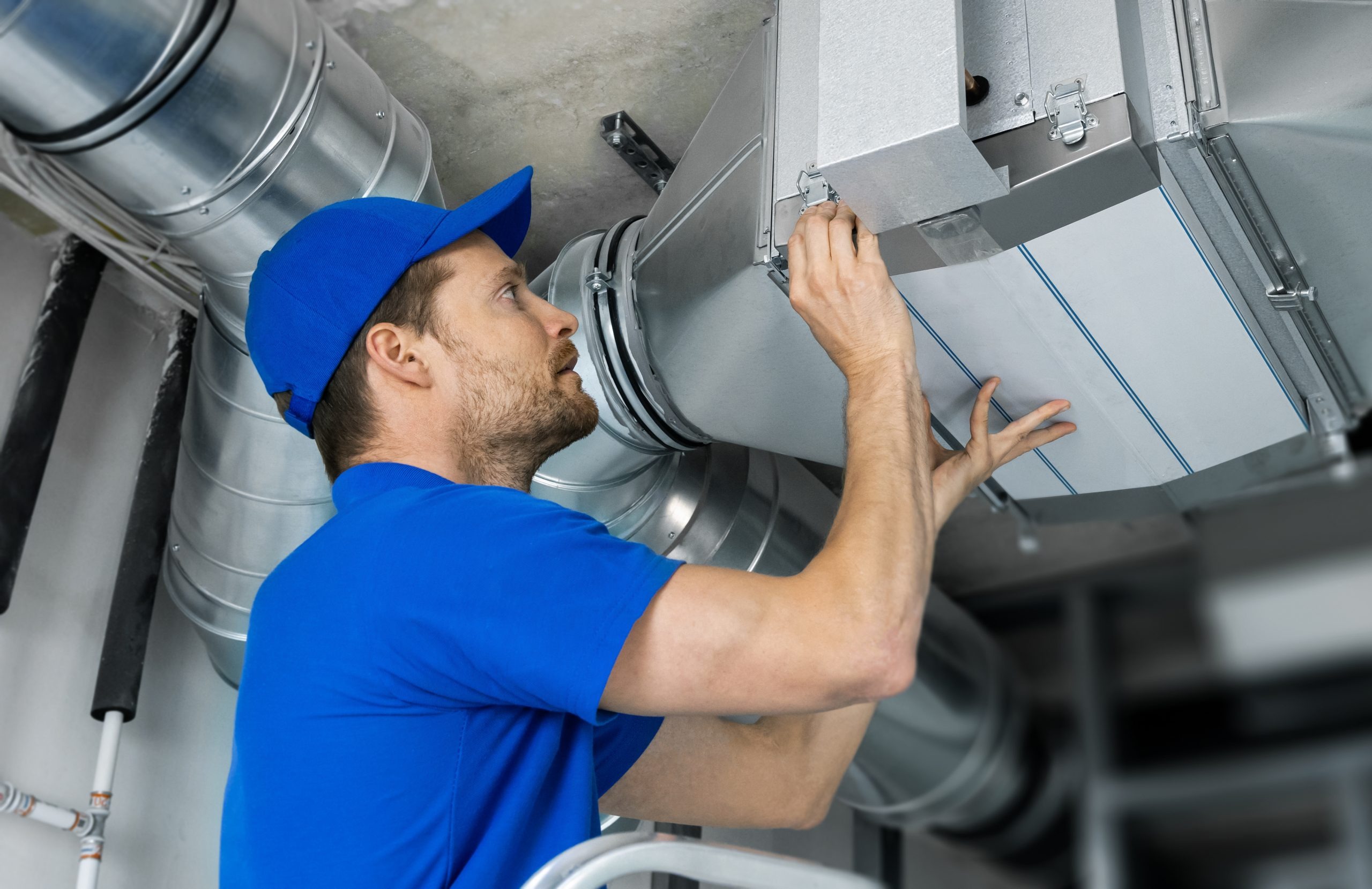 Technician in blue uniform and cap adjusting metal HVAC ductwork in a building ceiling.
