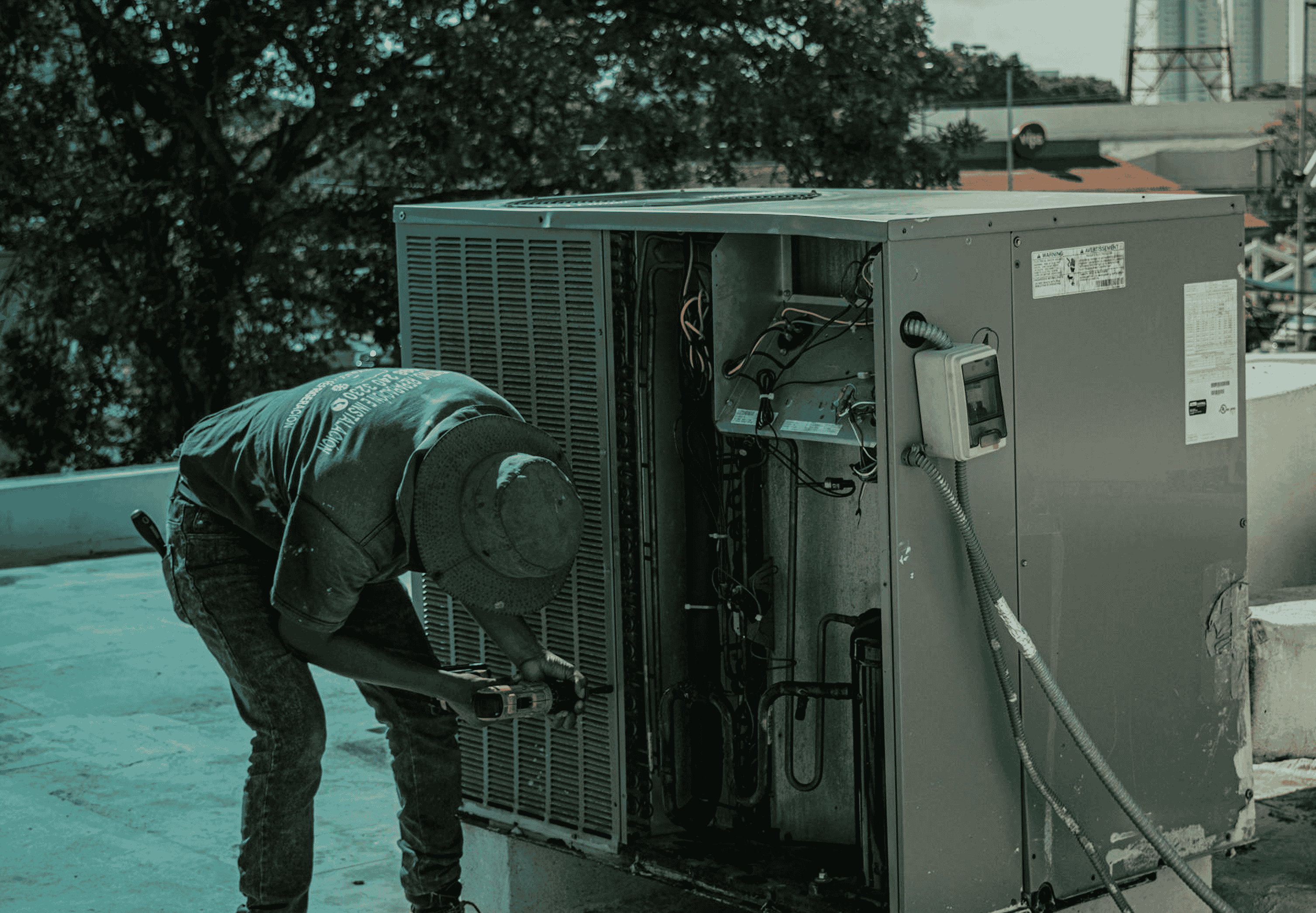 Technician wearing a hat using a power tool to repair an outdoor HVAC unit on a rooftop.