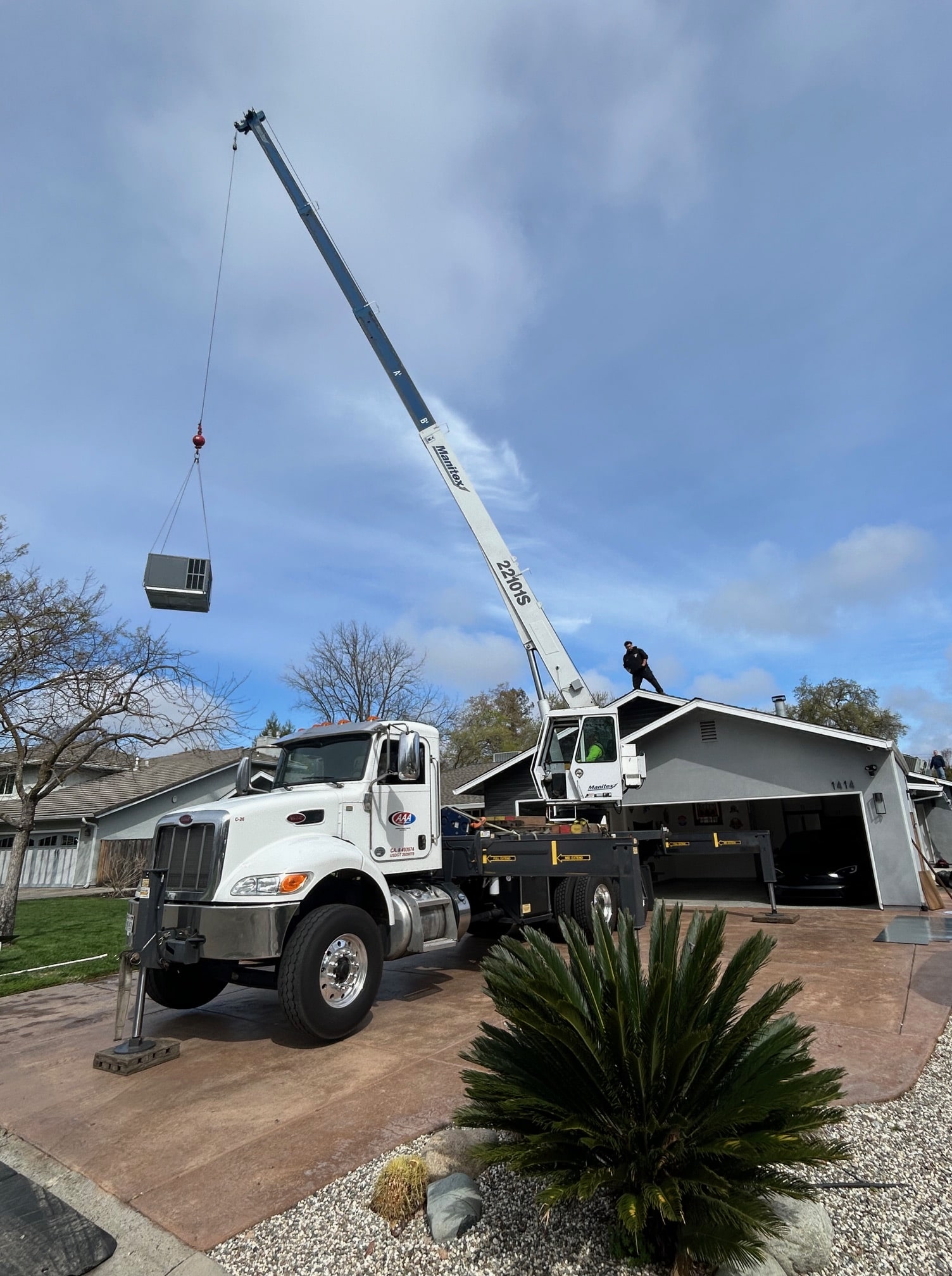 White crane truck with extended boom lifting an HVAC unit over a residential garage with a person standing on the roof.