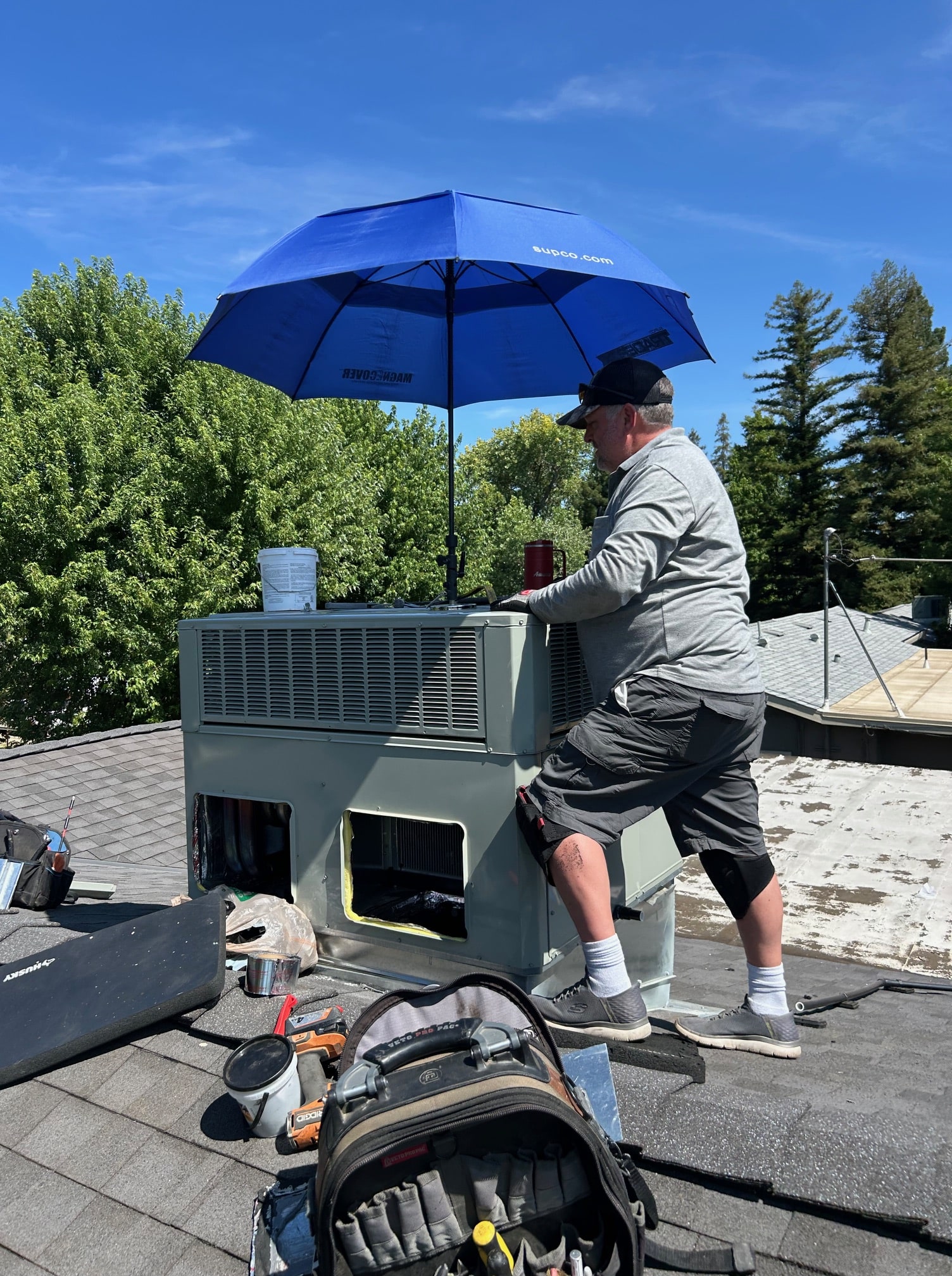 Technician wearing knee pads working on an HVAC unit on a rooftop under a blue umbrella on a sunny day.