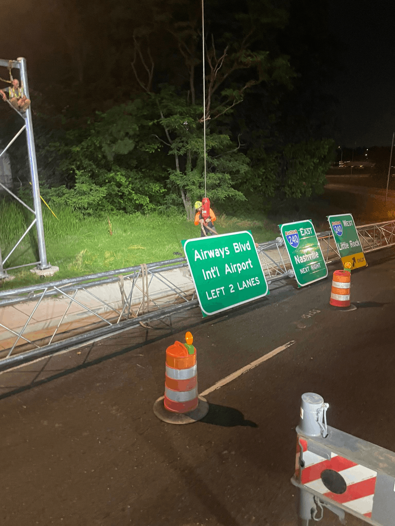 Nighttime road construction scene with worker in safety gear installing or repairing overhead highway signs for Airways Blvd, I-240 East to Nashville, and I-240 West to Little Rock.