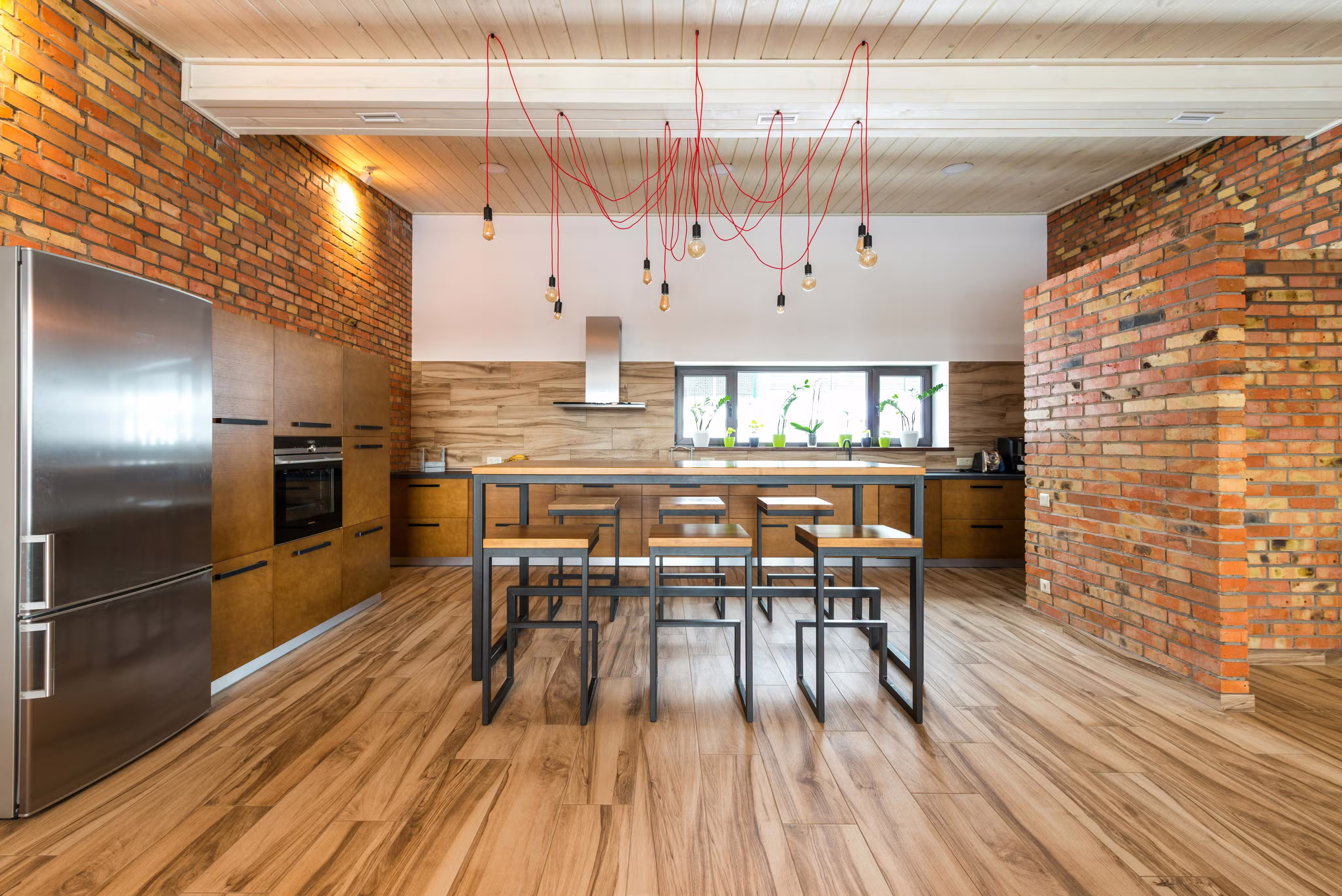Modern kitchen with wooden cabinets and floor, exposed brick walls, a stainless steel refrigerator, and a central wooden table with matching stools under hanging light bulbs with red cords.