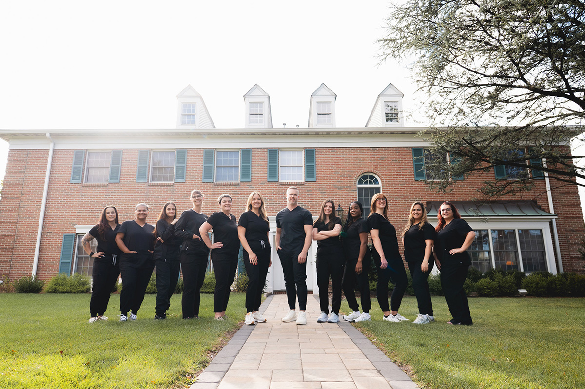 Group of twelve people dressed in black standing on a paved walkway in front of a large brick building with green shutters.