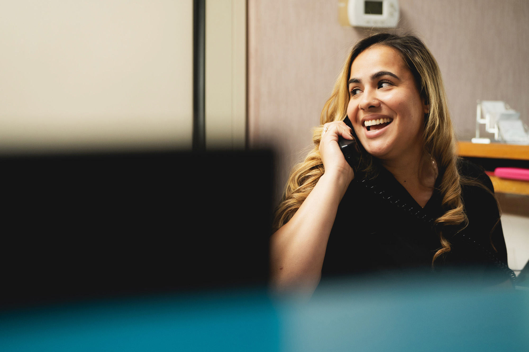 Smiling woman with long curly hair talking on a telephone in an office setting.