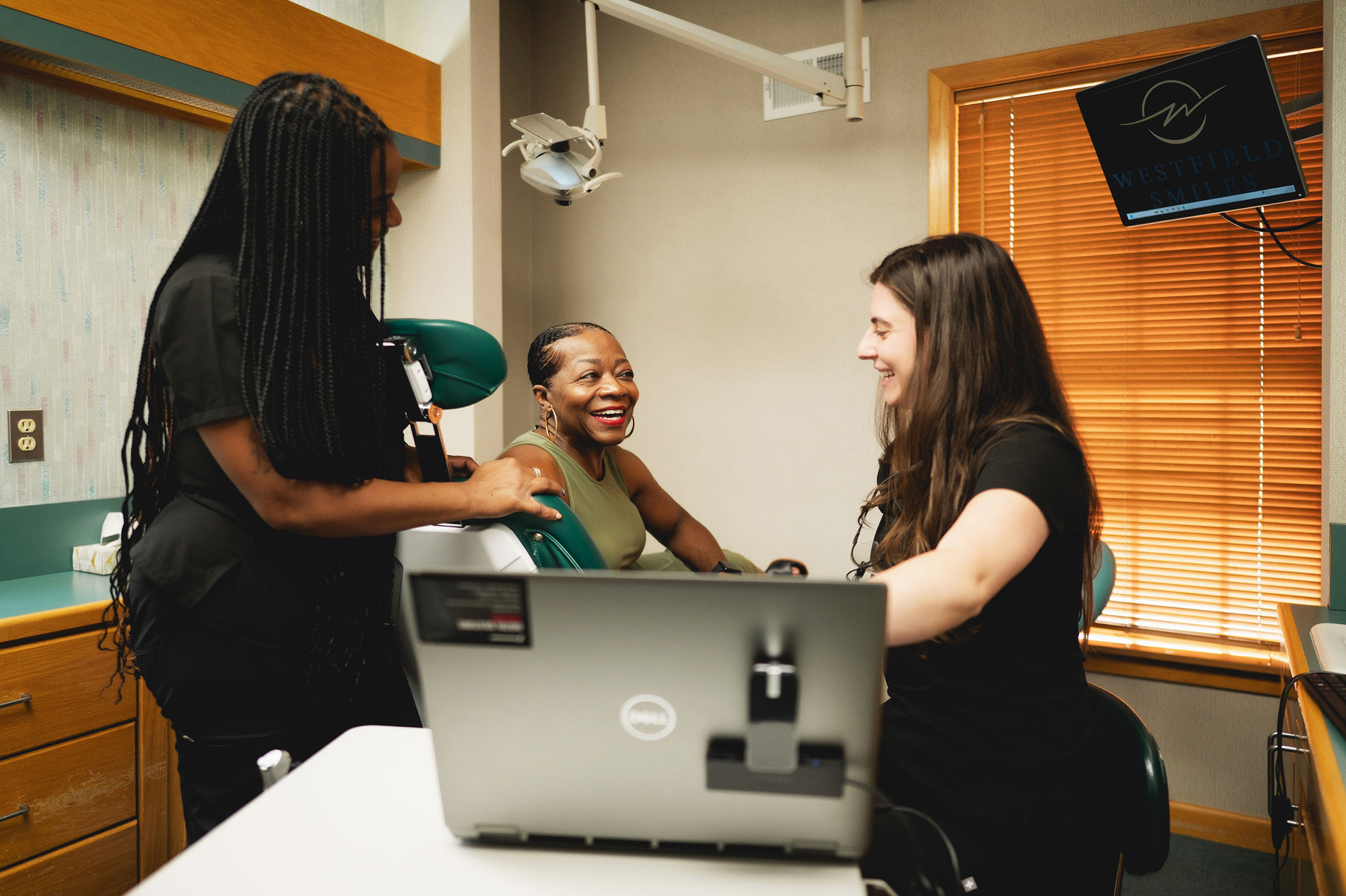 Two dental professionals engaging with a smiling female patient seated in a dental chair in a clinic room.