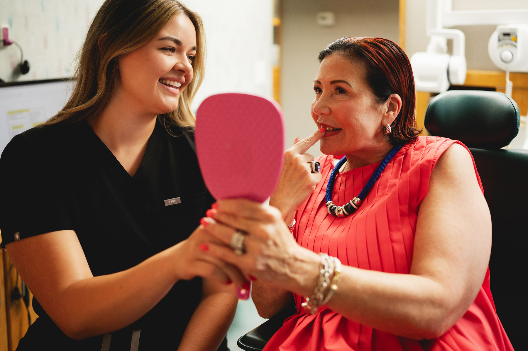 Dental professional in black scrubs smiling while holding a pink mirror for a seated woman in a red dress who is examining her teeth.