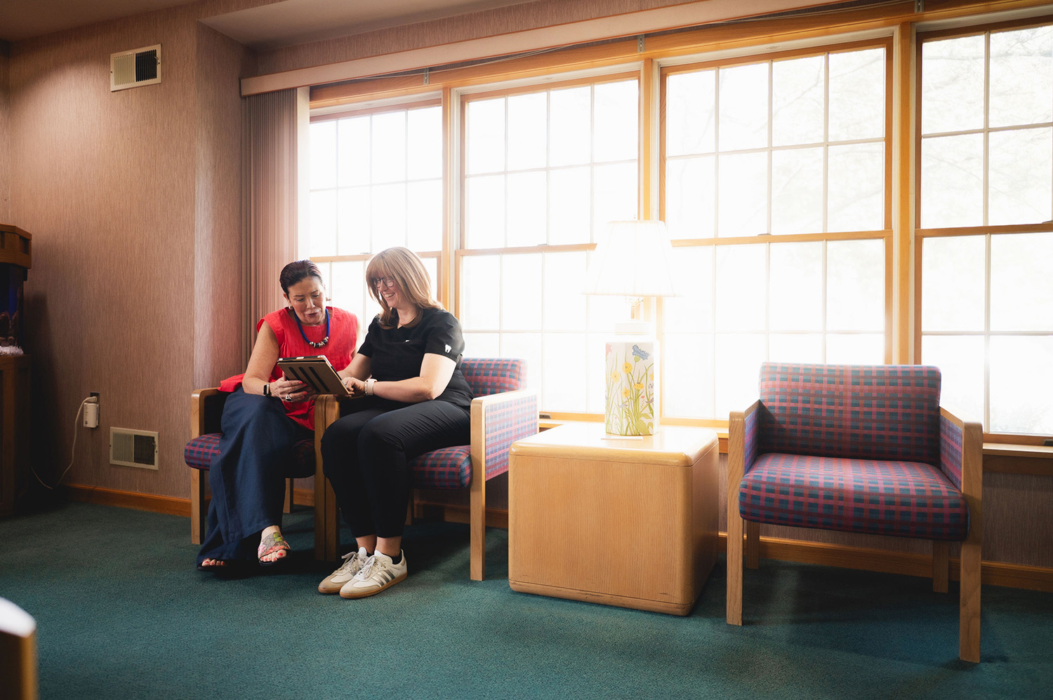 Two women sitting on patterned chairs in a bright room, looking at a tablet together and smiling.
