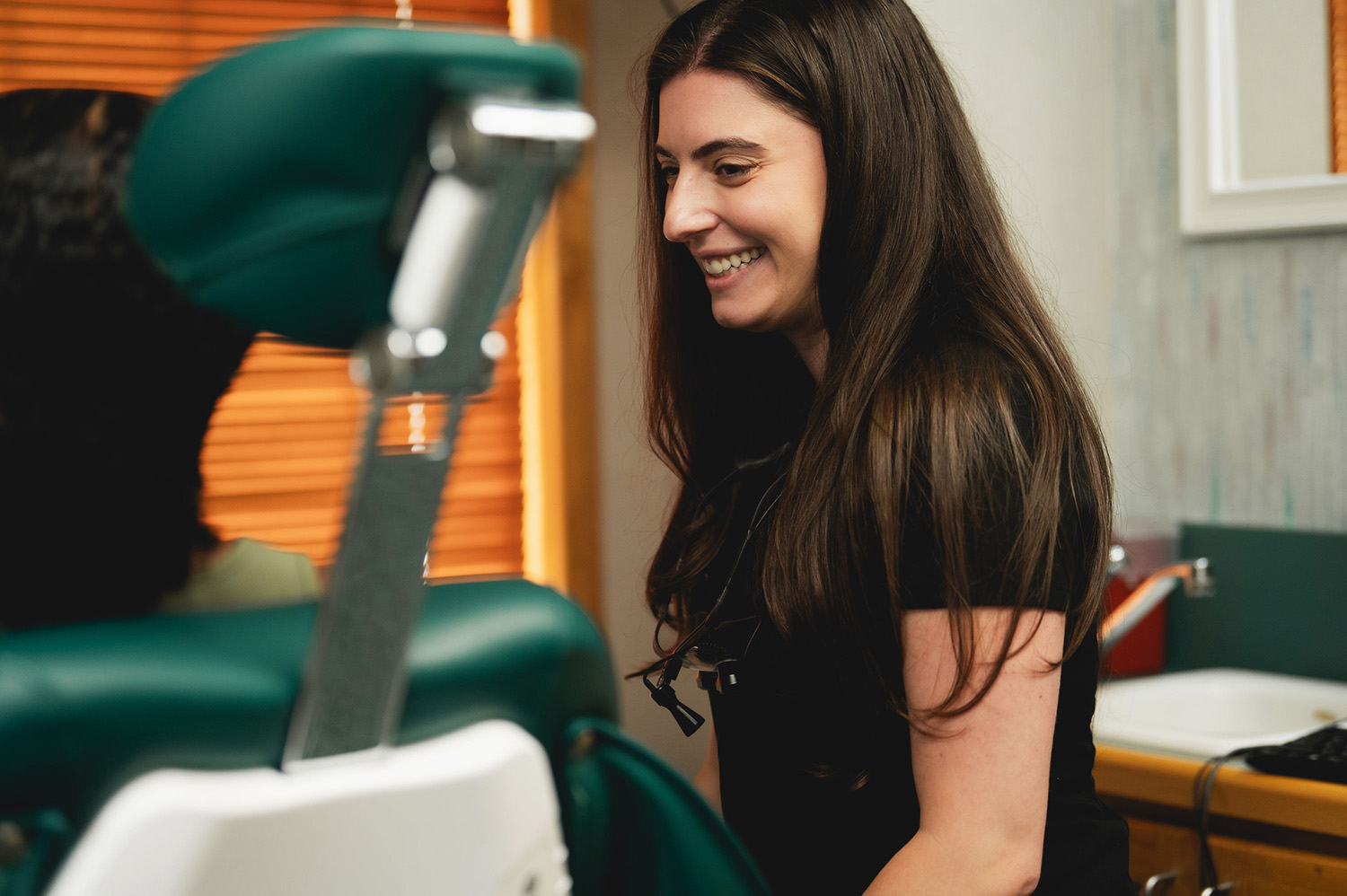 Smiling woman with long brown hair sitting at a green dental chair in a dental office.