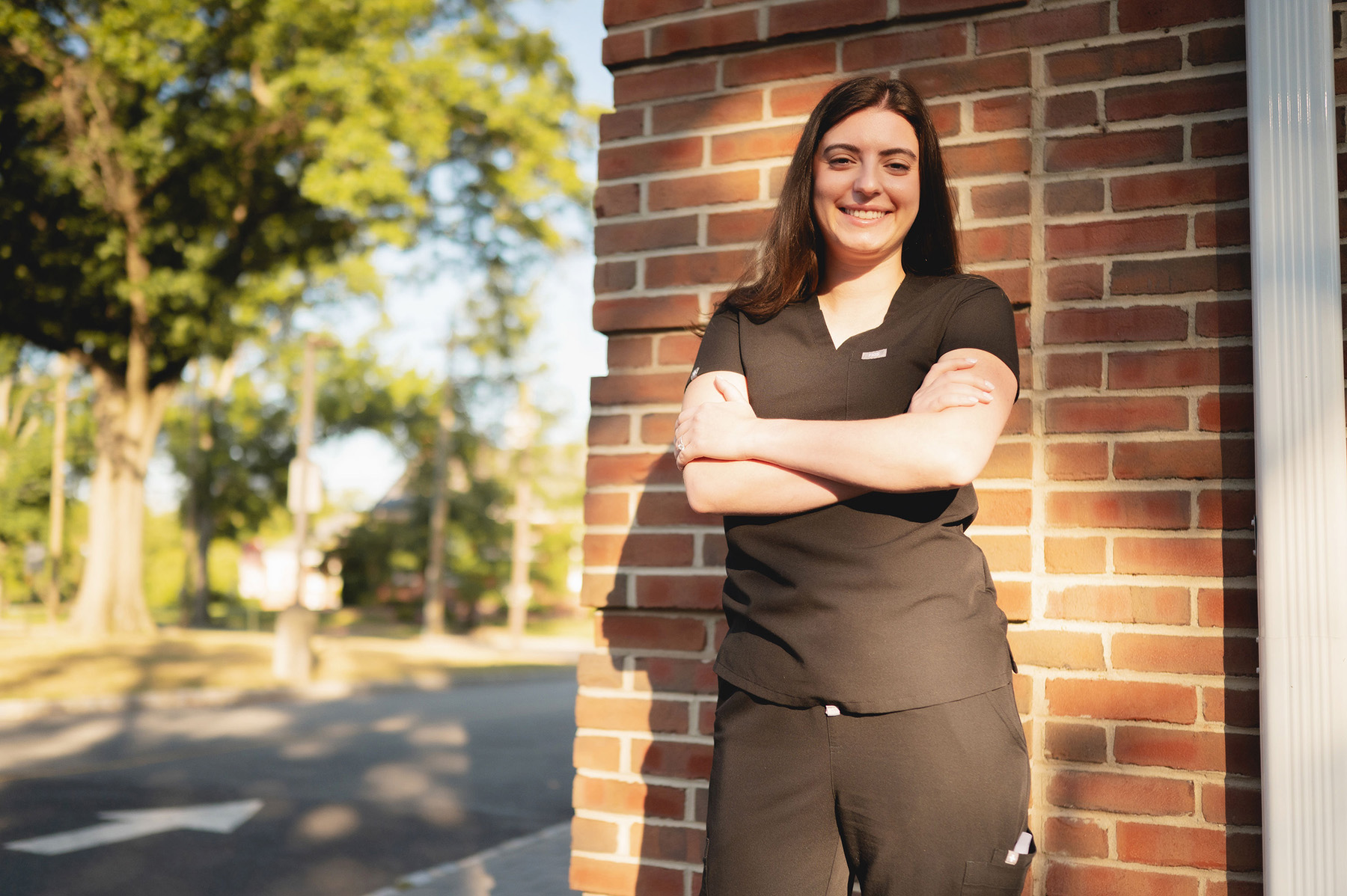 Smiling woman in black medical scrubs standing with arms crossed against a brick wall outdoors.