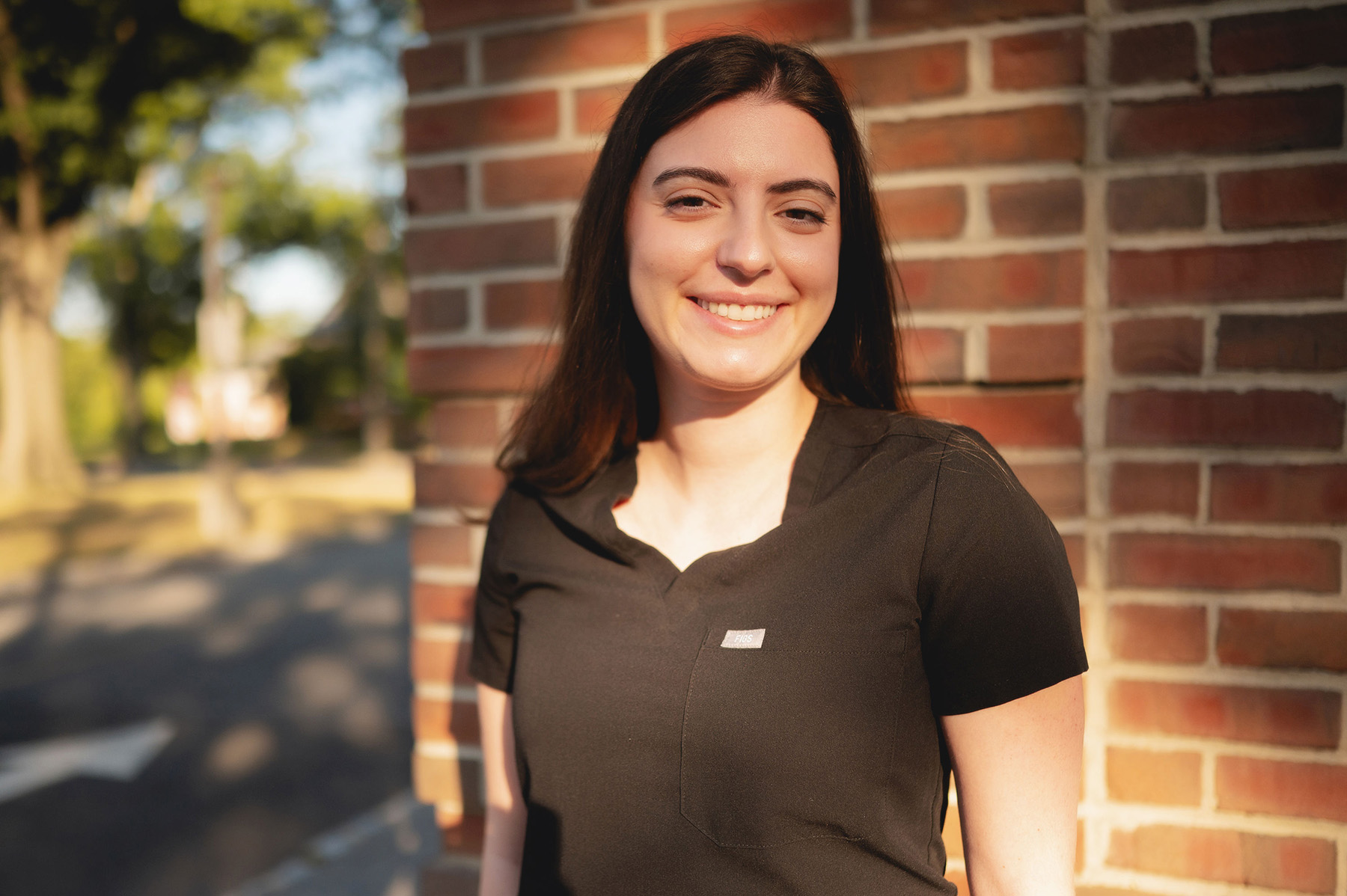 Smiling woman with long dark hair wearing a black top standing against a brick wall in natural sunlight.