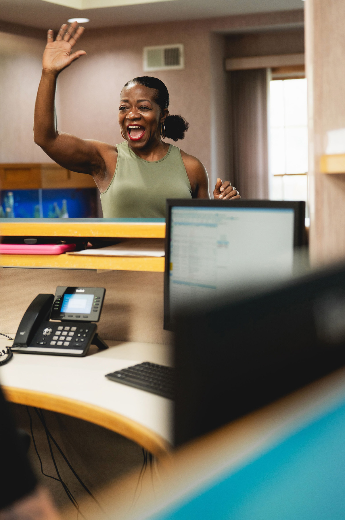 Smiling woman waving hello across an office reception desk.