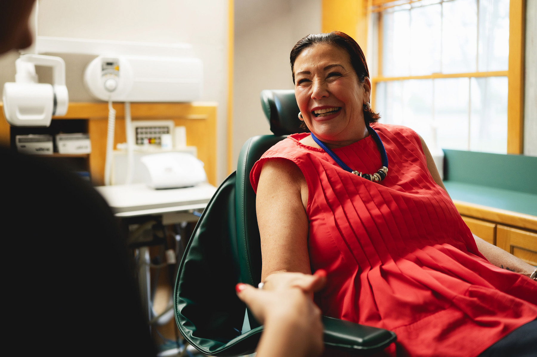 Smiling woman in a red top sitting in a dental chair shaking hands with someone.