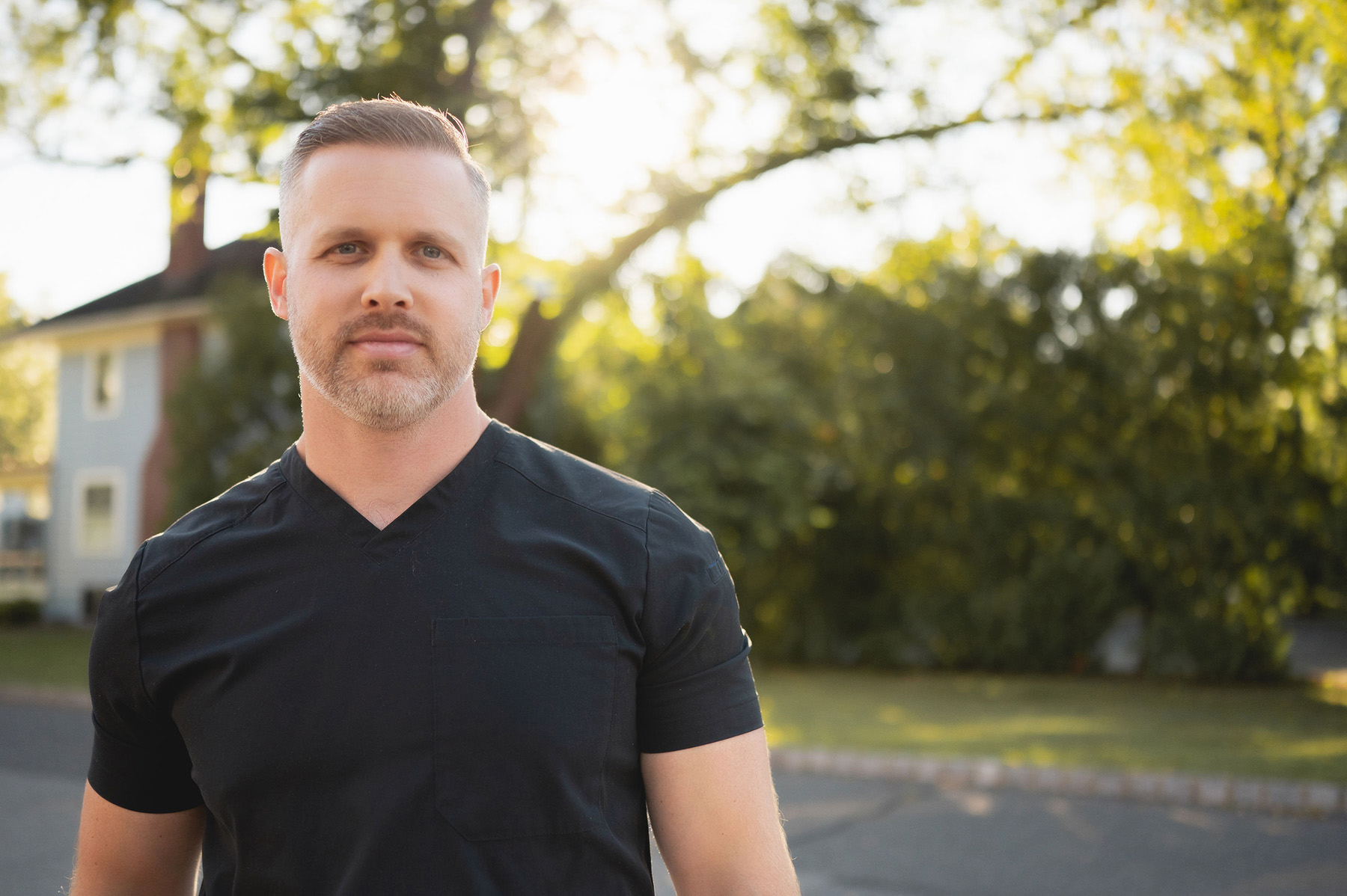 Man with light beard wearing black scrubs standing outside with trees and a house in the background at sunset.