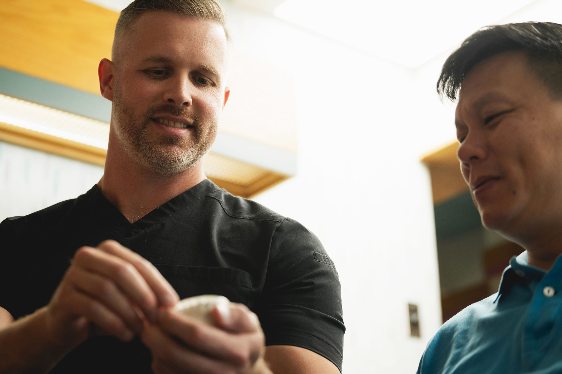 A male healthcare professional in black scrubs showing a dental model to a smiling male patient in a blue shirt.