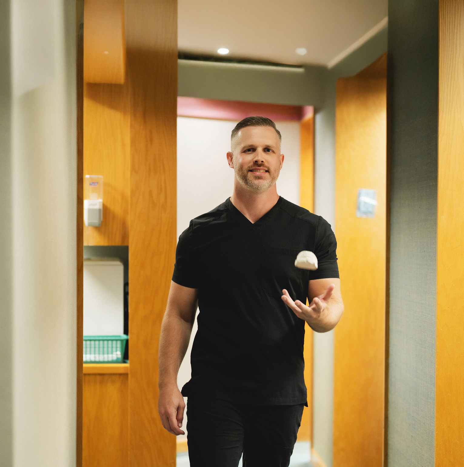 Man wearing black scrubs smiling and tossing a dental mold in a hallway with wooden panels.