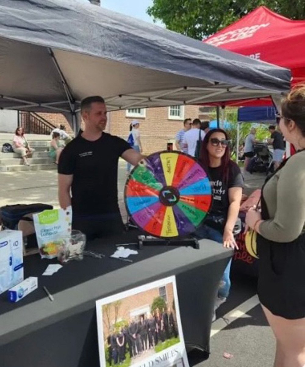Two people manning a booth with a colorful prize wheel under a canopy tent at an outdoor event, interacting with a visitor.