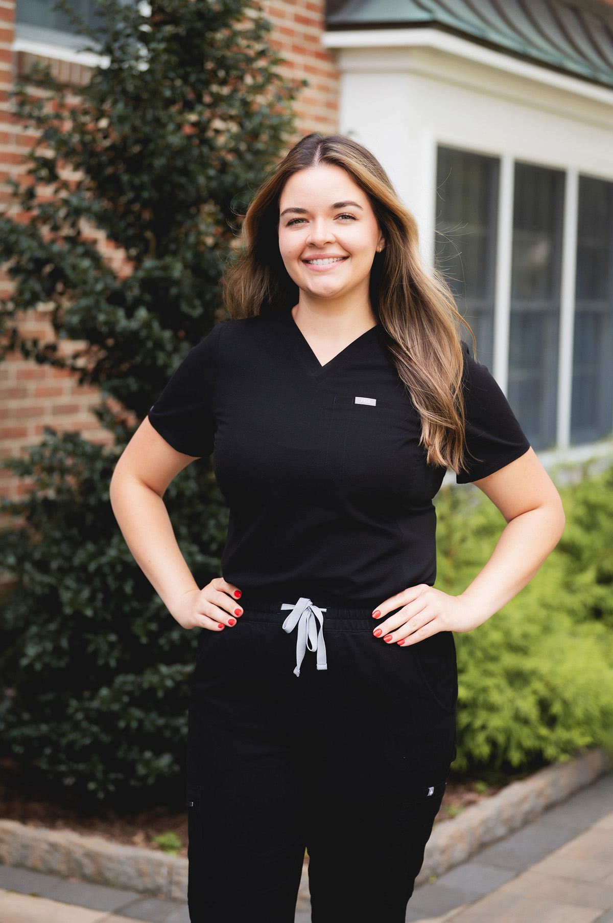 Smiling woman in black medical scrubs with hands on hips standing outdoors near greenery and a brick building.