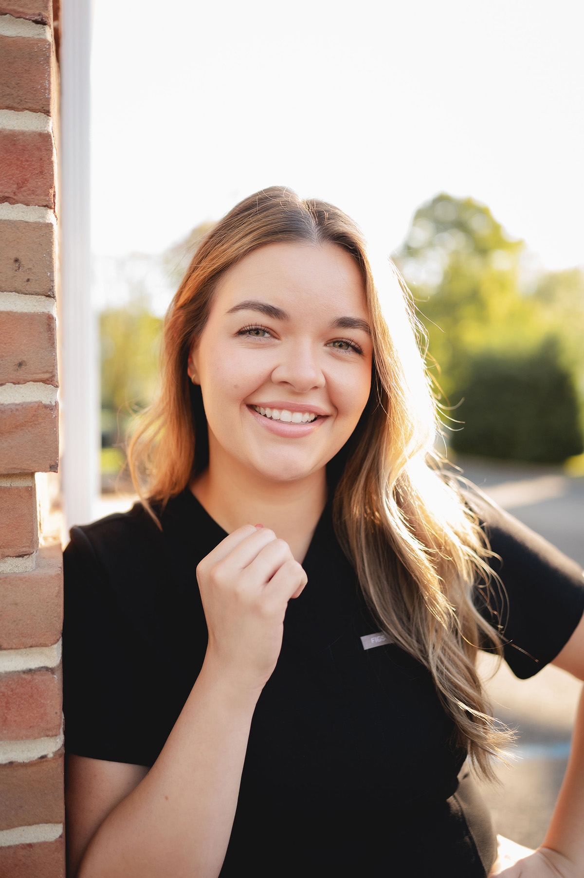 Smiling young woman with long hair leaning against a brick wall outdoors in sunlight.