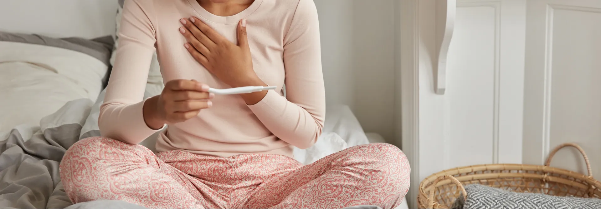 Woman sitting cross-legged on a bed holding a pregnancy test while touching her chest.