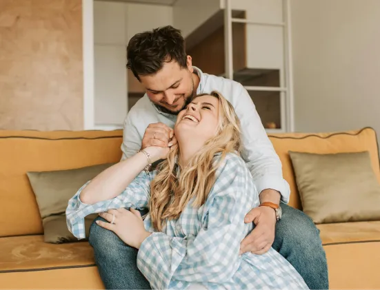 Smiling couple sitting on a yellow couch, the man affectionately hugging the woman who is laughing.
