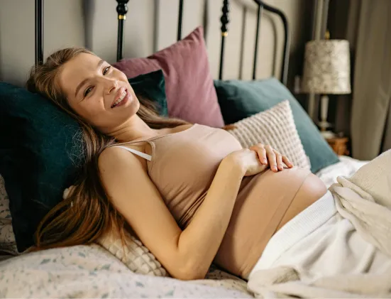 Pregnant woman lying on a bed, smiling and resting her hands on her belly surrounded by pillows.