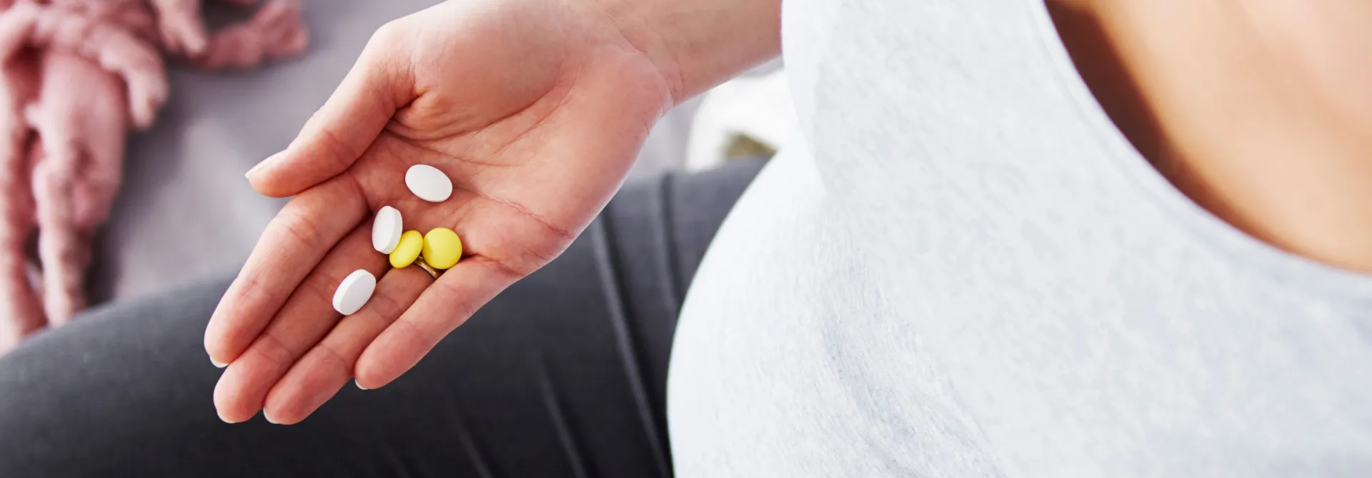 Close-up of a hand holding white and yellow pills resting on a pregnant belly.