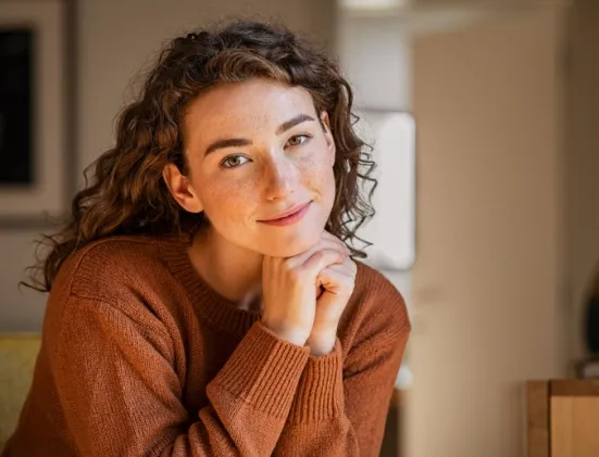 Smiling young woman with curly hair wearing an orange sweater, resting her chin on her hands in a cozy indoor setting.