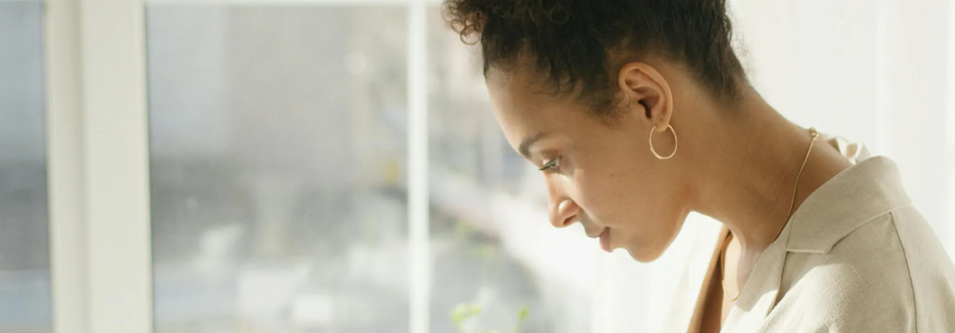 Profile of a woman with curly hair tied back, wearing a light shirt and hoop earrings, looking down thoughtfully.