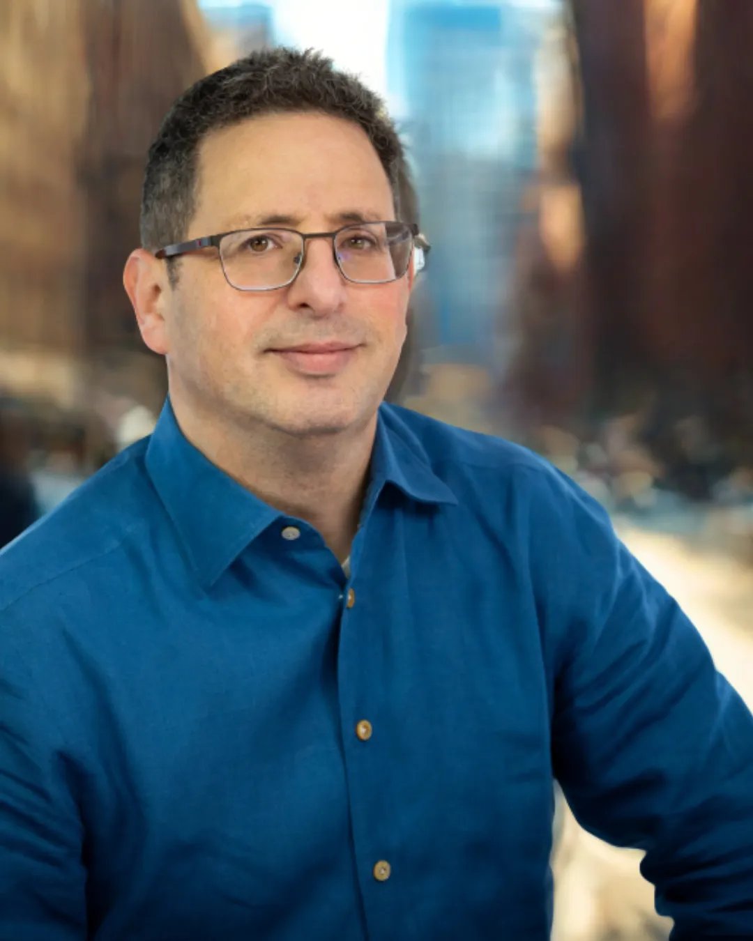 Man with short dark hair and glasses wearing a blue button-up shirt, standing outdoors with blurred city buildings in the background.