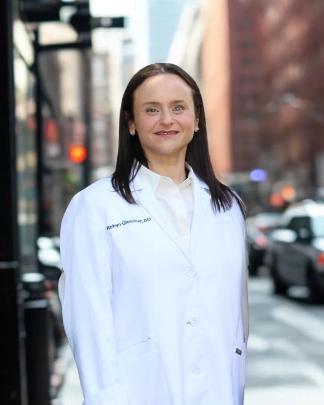 Robyn Glessner, DO, smiling in an embroidered white coat on a Manhattan street