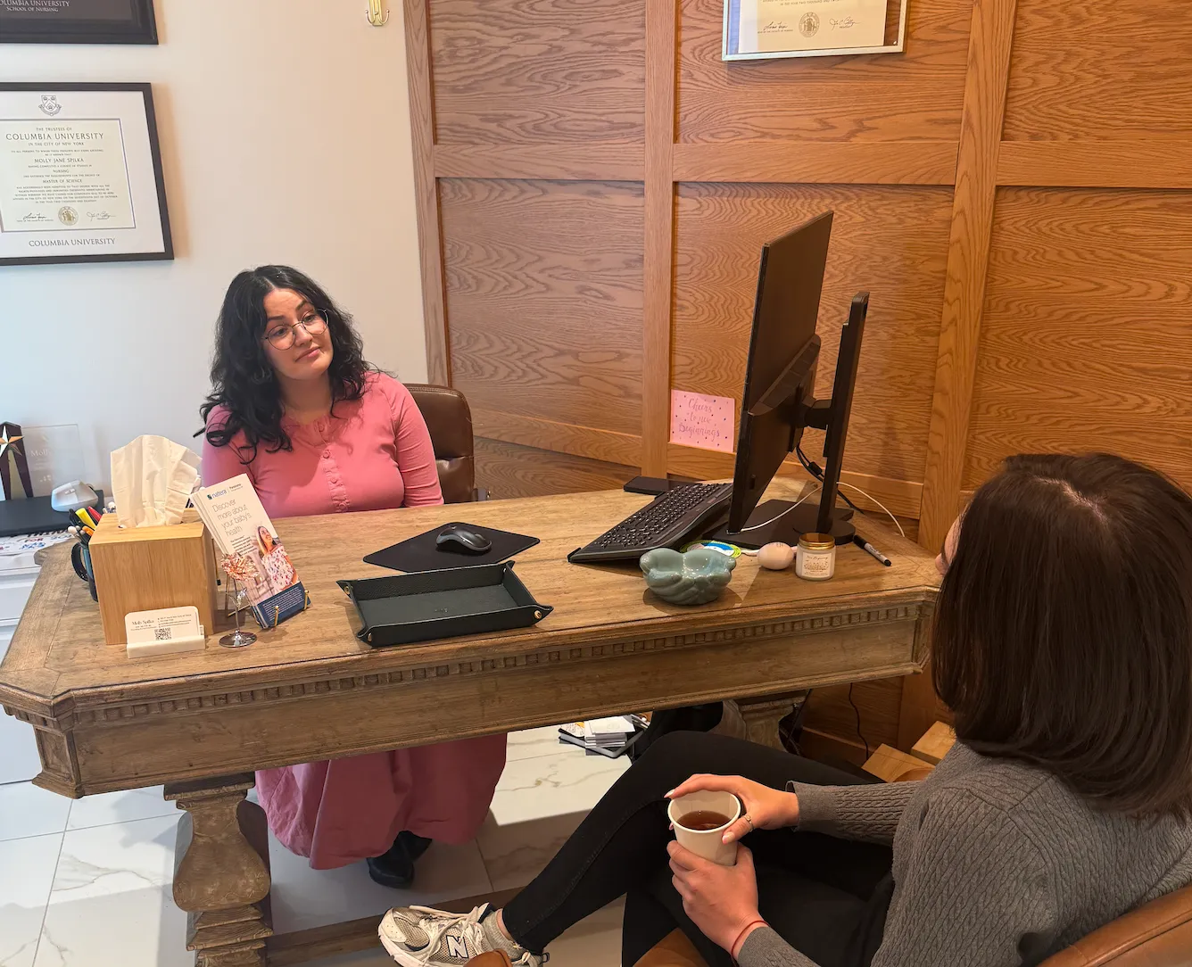 Provider in a pink dress chats across a wooden desk with a patient holding a mug, diplomas on the wall