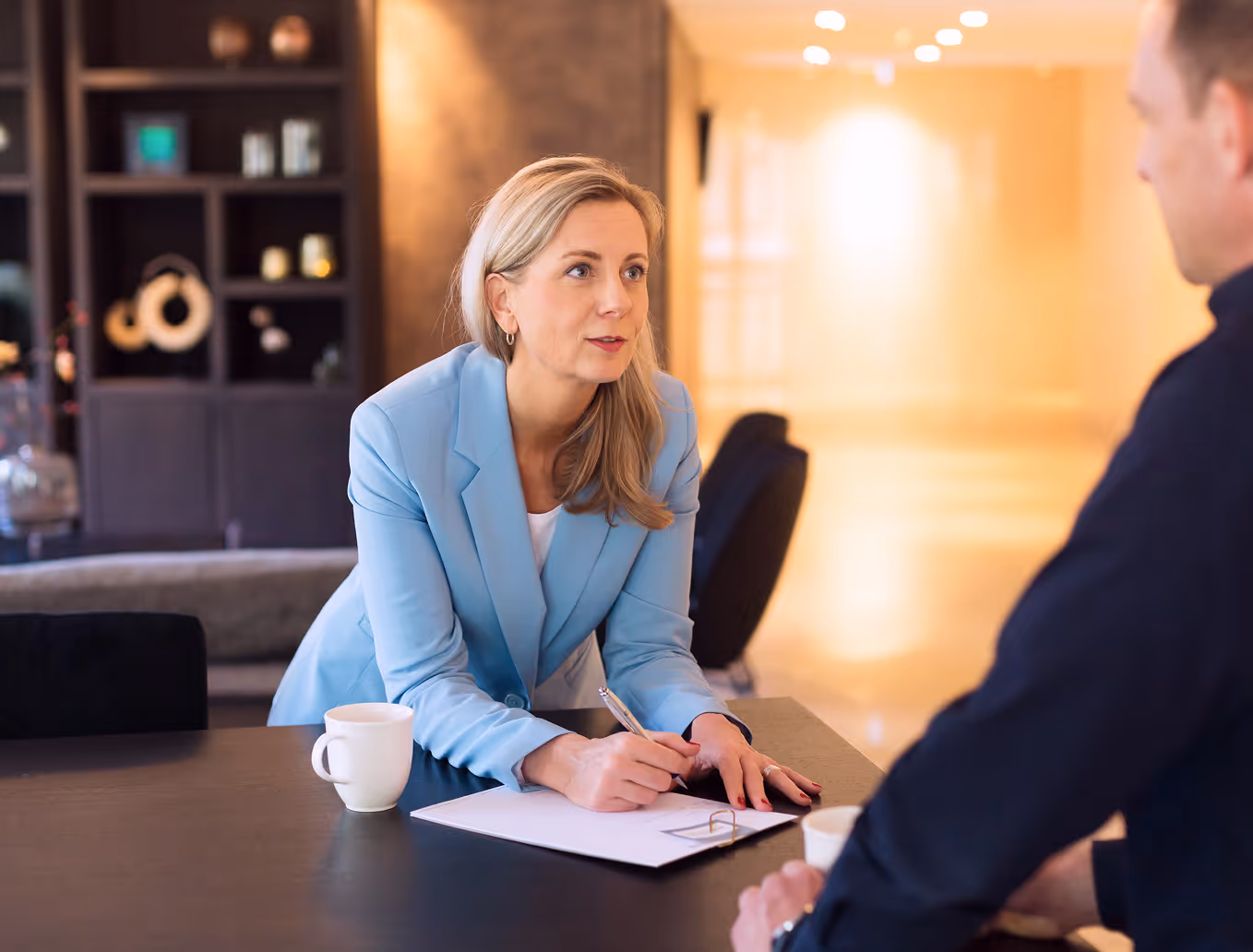 Woman in a blue blazer attentively speaking to a man across a table with a clipboard and coffee mugs in a modern office.