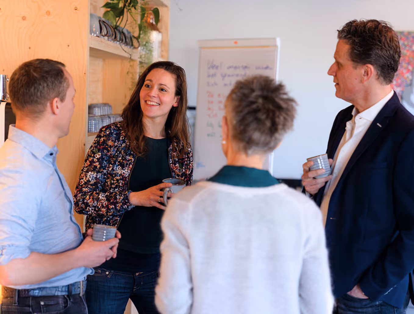 Four colleagues standing and chatting in a casual office setting, holding ceramic mugs.